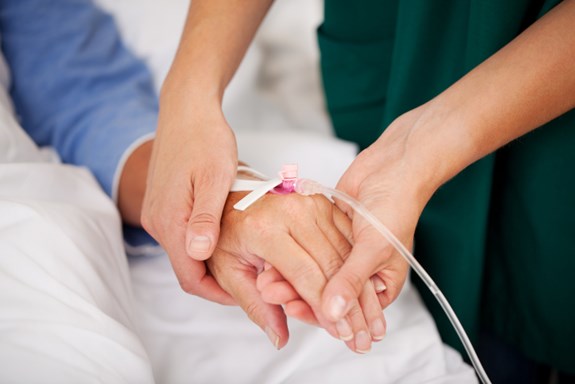 Fotografiet er eit nærbilde av tre hender. Ein sjukepleiar held ein pasient i henda. Pasienten har kanyle festa i handa. Foto: Mostphotos.