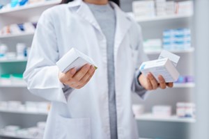 Bilde av en farmasøyt som holder i flere pakker med piller. I bakgrunnen er det en hylle med flere ulike typer medisiner.